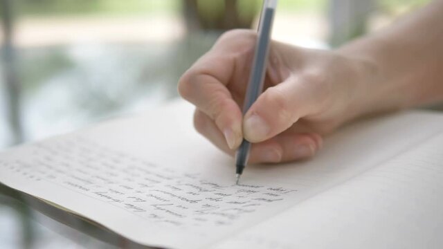 woman writes on open paper notebook page with black ball pen sitting at glass table in brightly lit restaurant extreme closeup