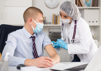 Doctor in face mask uses hypodermic needle and a syringe to put a shot of drug to patient in...