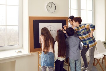 Modern school learning method for children education. Young caucasian female teacher teaching schoolchildren group reading clock time, minutes and hours. Tutor with students standing at class board