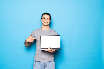 Young man holds the laptop pointing at the screen of the laptop smiling, on a blue background.