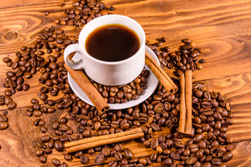 Cup of hot coffee, cinnamon sticks and scattered coffee beans on wooden table