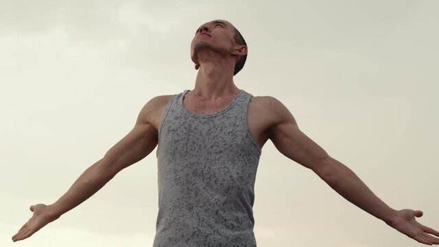 Man of athletic build in gray T-shirt is standing in outdoor against background of overcast sky, arms outstretched and looking up, enjoying rain, side and down view, slow motion