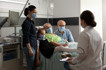 Naklejka premium Family visiting elderly senior woman patient while wearing protective face mask in hospital ward during clinical recovery. Practitioner doctor discussing disease symptoms explaining medical treatment