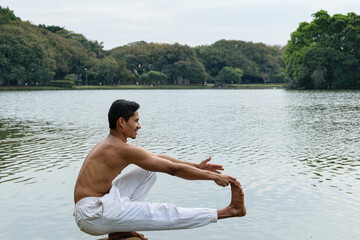 Brazilian young man crouched beside a lake, shirtless, stretching one of his legs and arms.