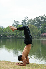 Young Brazilian man standing beside a lake, shirtless, doing yoga scorpion position.