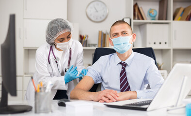 Doctor in white coat and protective mask giving vaccine injection to manager in office