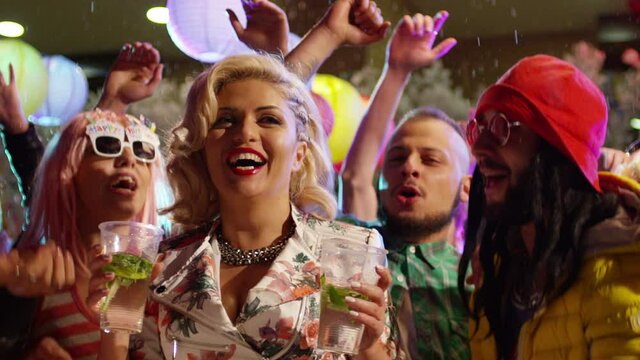 Blonde girl pouring two glass of alcohol over her head and mouth . Girl pours 2 cups of mojito over herself during colorful party . Crowd or young , stylish multi-ethnic people dancing . Slow Motion .