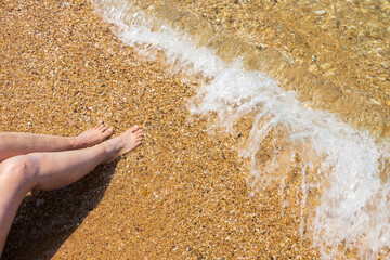 Beautiful view of female sitting on sand beach with legs in oncoming wave. Greece. 