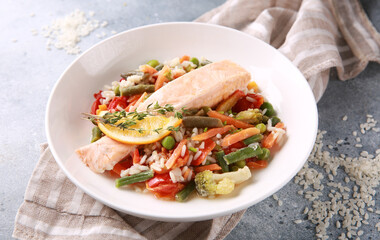 Seafood dish. Red fish with rice and vegetables. Salmon with lemon, beans, carrots, peas and cauliflower in a white plate on a gray table. Background image, copy space