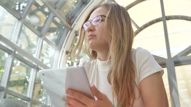 pretty long haired businesswoman in glasses works with contemporary tablet sitting at table in light cozy cafe low angle shot