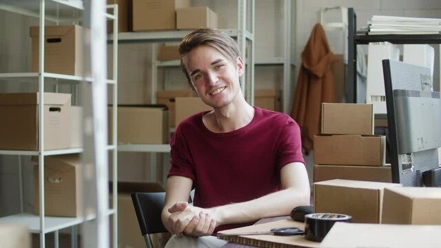 Happy worker of storage sitting and smiling, employee of warehouse, man looking in camera with smile on the background of cardboard boxes. Logistics, delivering and storage concept.