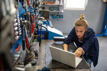 Mechanic using laptop at the repair garage. Young female mechanic with laptop. Business woman at a factory. Mechanic typing on a laptop at the repair garage
