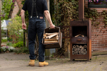 Man wearing casual clothes and suspenders carrying a big box with wooden logs to a metal wood stove in his garden while living the simple life in a rural area with greenery - Powered by Adobe