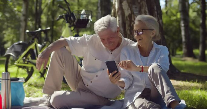 Happy mature couple relaxing in park on picnic using smartphone together