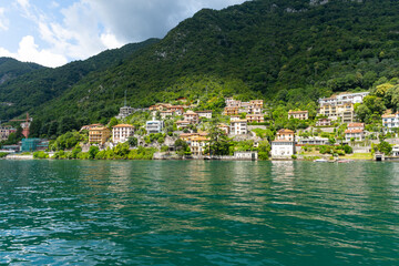 Fototapeta na wymiar Small town by Lake Como, seen from the lakeside. Beautiful village spread out over the hills, and all the way down to the lake. It is a beautiful sunny summer day