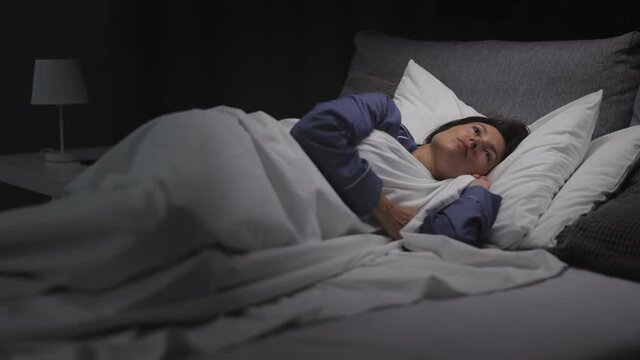 Displeased woman with dark hair turning around in bed because of insomnia. Caucasian lady in blue pajamas unable to sleep alone at home.