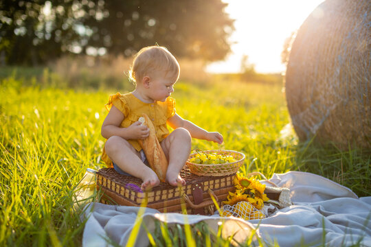 Little girl eats fresh baguette and fruit on a picnic at sunset lights in nature.