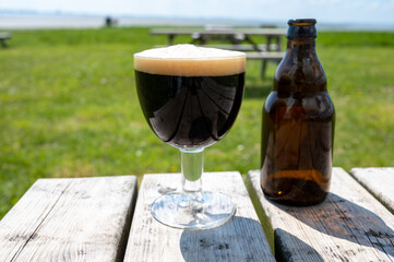 Glass of dark strong belgian beer served on outdoor terrace with green grass meadow on background