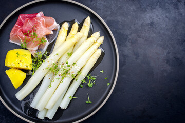 Traditional steamed white asparagus with cured ham and boiled potatoes garnished with butter sauce served as top view on a Nordic design plate with copy space right