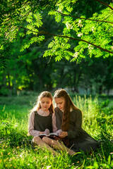 Two schoolgirls in school uniforms are reading a book while sitting on the green grass in the park. Do homework