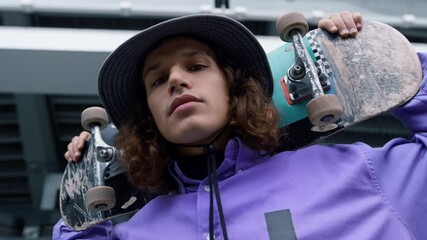 Stylish man looking camera outdoor. Skater holding skateboard on shoulders.