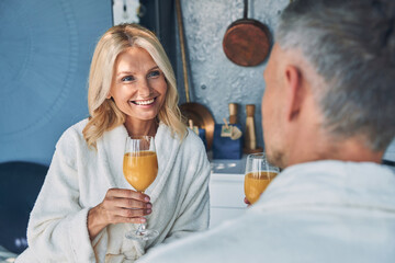 Beautiful mature couple in bathrobes enjoying fresh juice together while spending time in the domestic kitchen