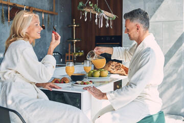 Fototapeta na wymiar Beautiful mature couple in bathrobes enjoying breakfast together while spending time in the domestic kitchen