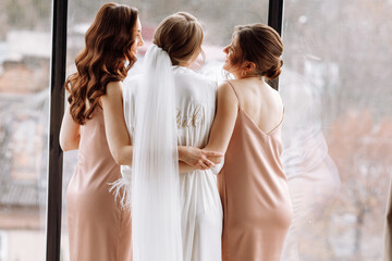 Fototapeta na wymiar Morning of bride. Gorgeous bride with best bridesmaids are having fun in hotel room near the large window. Sexy bridesmaids in exciting negligee. Wedding morning details. back view