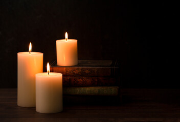 Obraz na płótnie Canvas Three Pillar Candles Burning in a Dark Room with a Stack of Antique Books