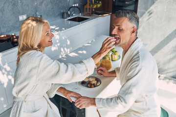 Top view of beautiful mature couple in bathrobes enjoying breakfast together while spending time in the domestic kitchen