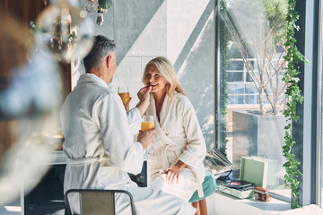 Beautiful mature couple in bathrobes enjoying breakfast together while spending time in the...