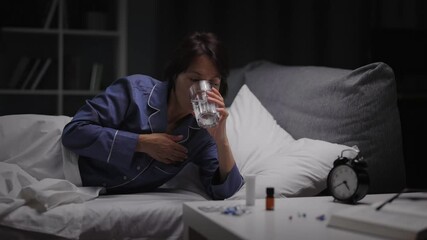 Caucasian middle aged woman in blue pajamas holding glass of water and pills while lying in bed. Tired brunette taking medicine from insomnia at night.