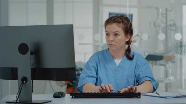 Stomatological assistant typing on computer keyboard sitting at oral clinic desk. Nurse using technology while professional orthodontist consulting patient with equipment in background