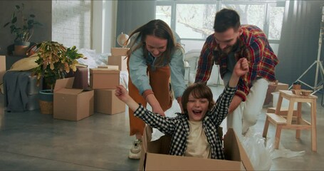 Happy parents have fun pushing excited cute boy in cardboard box on the floor celebrating moving in new home slow motion