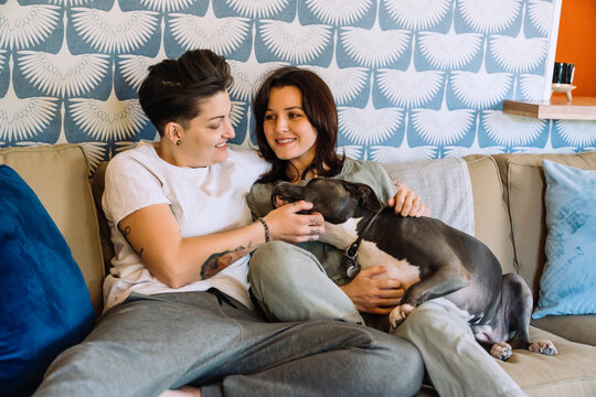 Happy Lesbian Couple Cuddling On Couch With Pet Dog