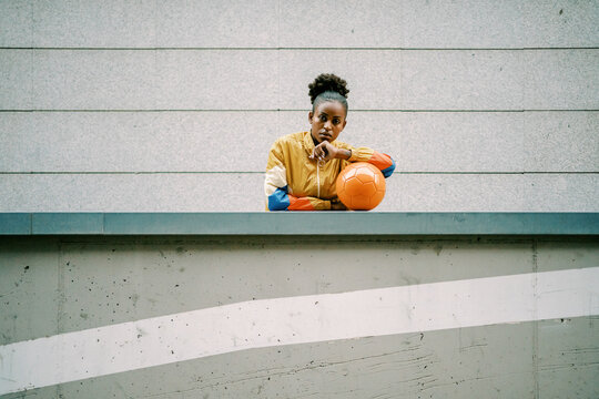 Serious female footballer standing with ball in street - Powered by Adobe