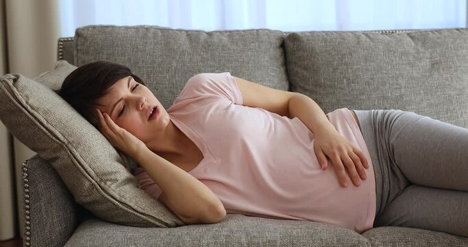Unhappy tired young pregnant woman suffering from headache or negative thoughts, feeling fear of childbirth, worrying about bad prenatal screening tests results, resting lying on comfortable sofa.