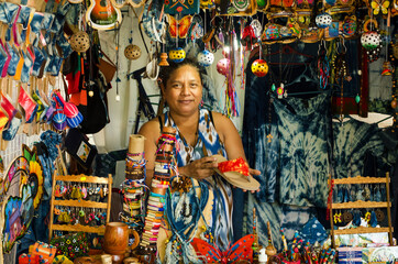 Tanned woman smaling  showing a shoe in her little store surrounded by colorful hand craft...
