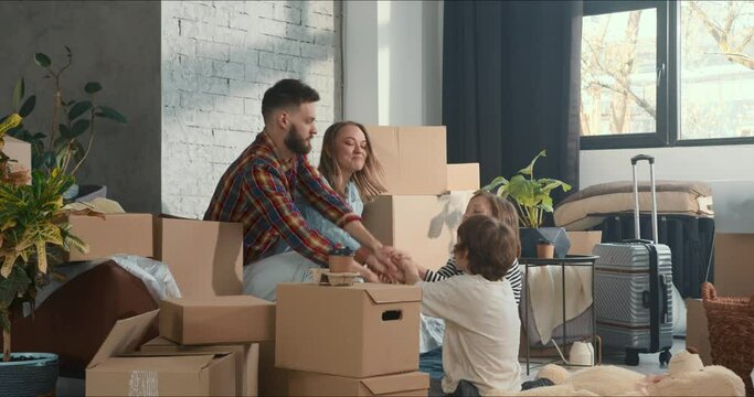Reaching life goals together. Young happy family with two kids join hands among boxes in light apartment after moving in