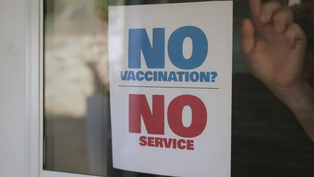 A small business owner places a NO VACCINATION NO SERVICE sign on his front door window. Some businesses started to customers to show proof of their coronavirus vaccination before entering.  	