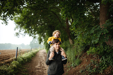 father walking on a dirt road with daughter in his neck 