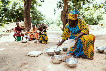 Colourfully dressed black African mother preparing food for her family, with her daughters waiting in the background