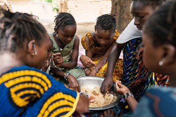 Group of coulorfully dressed black girls sitting around a big metal bowl, sharing a typical African...