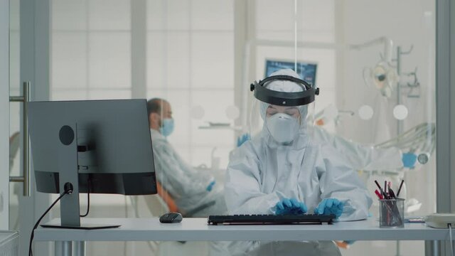 Portrait of dental assistent typing on computer keyboard wearing protection uniform with face shield, mask, gloves and coverall. Stomatology nurse using monitor technology at oral clinic