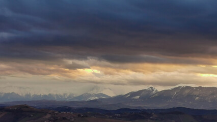 Obraz na płótnie Canvas Montagne innevate dell’Appennino al tramonto e grandi nuvole colarate di arancio nel cielo