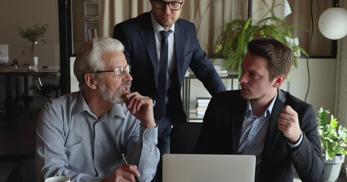 Older and younger male office employees gather together using laptop working on common project discuss collaborative corporate task share ideas and knowledge provide assistance during group meeting.