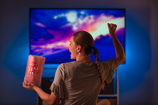 Evening. Rest. A young blonde woman is watching her favorite movie on TV. She reacts emotionally, waves her hand and smiles. In her other hand, she has a pack of popcorn.
