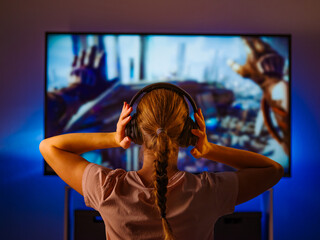 Video games. Video industry. Sports betting. A young blonde girl shooter in headphones sits in front of a large computer monitor and plays a video game. Shooting from the back.