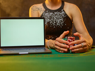 A young woman in an evening dress is gambling in an online casino. There is a laptop on a green poker table. Woman hands holding colored chips. Gambling. Casino. Night club. Online casino..