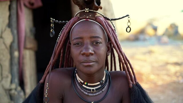 Married Himba woman looking at camera wearing traditional jewelry and Erembe headpiece at her village near Kamanjab in Namibia, Africa, slow motion shot.